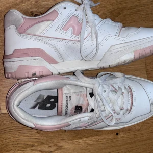 New Balance 550 sneakers i vitt och rosa - Snygga New Balance 550 sneakers i vitt med rosa detaljer. Skorna har snörning och en rund tå, vilket ger en klassisk look. Perfekta för en stilren och sportig stil.