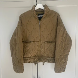 Grön quiltad jacka från Zara - Säljer en grön quiltad jacka från Zara med dragkedja framtill. Jackan har en unik vågig quiltad design och långa ärmar. Perfekt för kyligare dagar.