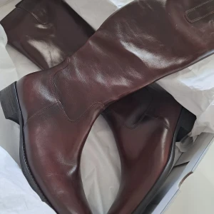 Bruna skinnstövlar från Gabor - Snygga bruna stövlar i skinn från Gabor. De har en klassisk design med rund tå och låg klack, perfekt för en stilren look. Stövlarna är knähöga och har en dragkedja på insidan för enkel på- och avtagning.