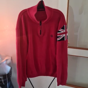 Röd stickad tröja med Union Jack-motiv - Säljer en röd stickad tröja med en dragkedja vid halsen och ett Union Jack-motiv på ärmen. Tröjan har långa ärmar och ribbade muddar. Perfekt för en cool och avslappnad stil.