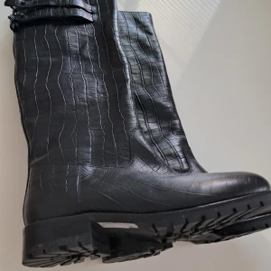 Svarta krokodilpräglade stövlar från Cosmo Paris - Snygga svarta stövlar med krokodilpräglat mönster och justerbara spännen upptill från Cosmoparis. Perfekta för en stilren look med robust sula för extra grepp. Storlek 40.