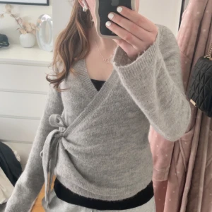 Grå omlottkofta från Gina tricot - Säljer en mysig grå omlottkofta med knytband i midjan i stolek M.  Koftan är i väldigt fint skick! Inga hål, fläckar och inget har gått av.