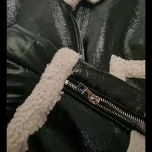 Svart jacka med teddyfoder från FB Sister - Snygg svart jacka från FB Sister med glansig yta och mysigt vitt teddyfoder. Jackan har dragkedja och dekorativa detaljer i teddy på krage och ärmar. Perfekt för kyligare dagar!