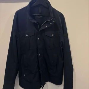 Marin blå jacka från J.Lindeberg - Snygg marin blå jacka från J.Lindeberg med dragkedja och knappar framtill. Jackan har två bröstfickor med lock och knappar samt en hög krage. Perfekt för en stilren look.