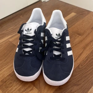 Adidas Gazelle sneakers i mörkblå mocka - Snygga Adidas Gazelle sneakers i mörkblå mocka med vita detaljer och klassiska tre ränder på sidan. Skorna har snörning och en vit sula som ger en stilren look. Använda en gång hemma men de passade inte! STORLEK: 37 och en halv💕