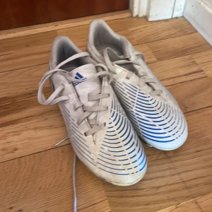 Vita och blå fotbollsskor från Adidas - Snygga Adidas Predator fotbollsskor i vitt med blå detaljer och snörning. Perfekta för fotbollsplanen med sin stilrena design och funktionella dobbar.