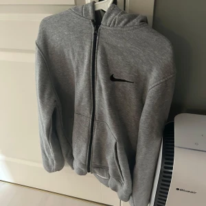 Grå hoodie från Nike - Säljer en stilren grå hoodie från Nike med dragkedja och huva. Den har en liten svart Nike-logga på bröstet och är perfekt för en avslappnad stil. Passar bra till både träning och vardag.