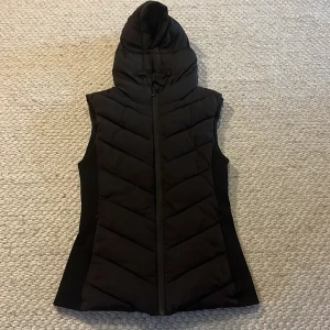 Svart dunväst från Zara - Snygg svart dunväst från Zara med huva och dragkedja. Perfekt för lager-på-lager under kyliga dagar. Västen har en stilren design med quiltade detaljer som ger en modern look. Knappt andvänd.