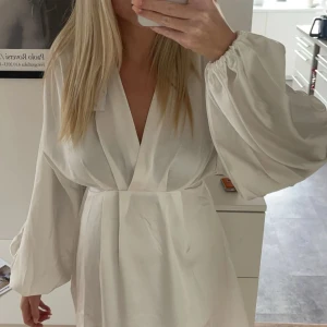 Vit långärmad klänning - Elegant vit klänning med v-ringning och långa puffiga ärmar. Klänningen har en omlott-design som ger en stilren look. Perfekt för en chic och sofistikerad stil.