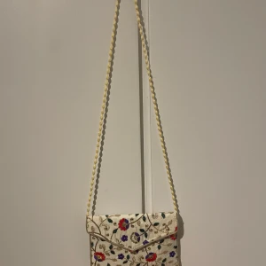 Broderad axelväska med blommönster - Charmig axelväska i beige med vackert broderade blommor i rött, lila och guld. Väskan har en gräddvit axelrem. Perfekt för att ge en färgglad touch till din outfit.