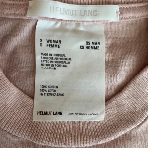 Ljusrosa t-shirt från Helmut Lang - Säljer en stilren ljusrosa t-shirt från Helmut Lang med tryck på framsidan. Tillverkad i 100% bomull, vilket gör den mjuk och bekväm. Perfekt för en avslappnad look. Liten fläck på magen 