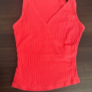 Röd ribbad omlottopp från Bershka - Säljer en snygg röd ribbad omlottopp från Bershka. Toppen är ärmlös och har en stilren omlottdesign som ger en modern look. Perfekt för att lyfta vilken outfit som helst. Passar bra till både jeans och kjol!