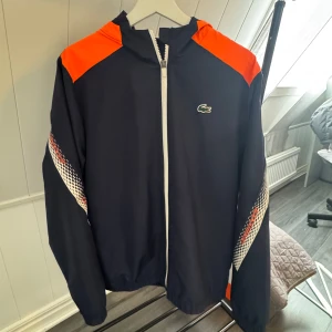 Mörkblå vindjacka från Lacoste - Snygg mörkblå vindjacka från Lacoste med orange detaljer på axlarna och ett coolt mönster längs ärmarna. Jackan har dragkedja framtill och en klassisk Lacoste-logga på bröstet. Perfekt för en sportig look! Endast använd 2 gånger!