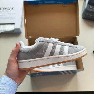 Adidas Campus 00s 37 2/3 - Snygga grå sneakers från Adidas med klassiska vita ränder och en gummisula i kontrasterande färg. Skorna har snörning och är tillverkade i mocka för en stilren look. Perfekta för en avslappnad stil.