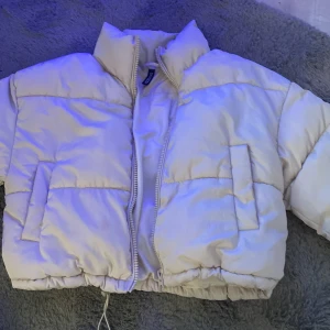 Vit pufferjacka från Divided - Säljer en snygg vit pufferjacka från Divided. Jackan har en dragkedja framtill och är perfekt för kyligare dagar. Den har en hög krage och två praktiska fickor på sidorna. Perfekt för att hålla värmen med stil!