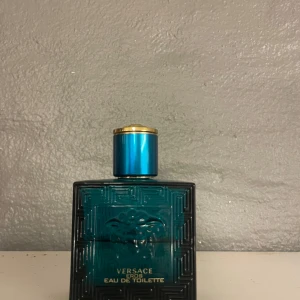 Versace Eros Eau de Toilette herrparfym - Versace Eros Eau de Toilette för herr, med en stilren blå glasflaska och guldiga detaljer på korken. Flaskan har ett klassiskt Versace-mönster och Medusa-symbolen framtill. Doften är fräsch och maskulin.
