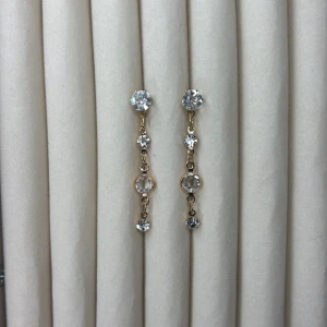 Guldiga örhängen med kristaller - Säljer ett par eleganta guldiga örhängen med fyra gnistrande kristaller i rad. De har en tunn kedja mellan stenarna och ger ett stilrent och lyxigt intryck. Perfekta för att lyfta din look! Finns matchande armband. 