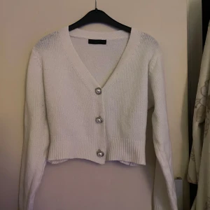 Vit croppad kofta med pärlknappar - Superfin vit stickad kofta med v-ringning och tre dekorativa pärlknappar framtill. Modellen är croppad och har långa ärmar samt bred ribbad mudd nedtill och vid ärmslut. Perfekt att slänga över en topp för en söt look.