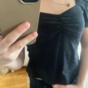 Svart blus med puffärm och rynk framtill - Säljer en svart blus med kort puffärm och rynkdetalj vid bysten. Blusen har fyrkantig ringning och är tillverkad i ett lätt, mönstrat tyg. Perfekt för dig som vill ha en stilren men ändå söt topp.