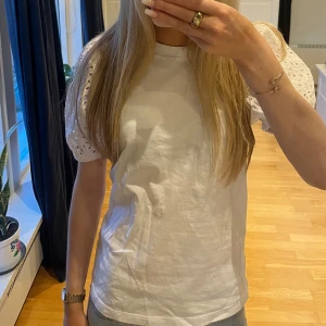 Vit t-shirt med spetsärmar - Säljer en vit t-shirt med korta ärmar i spets och broderade detaljer. T-shirten har en klassisk rund halsringning och är i en rak passform. Perfekt för dig som vill ha en basic t-shirt med en twist!