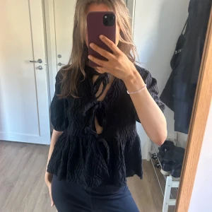 Svart knytblus med struktur - Säljer en svart blus med knytning framtill och krinklad struktur. Blusen har korta ärmar och en lös passform. Andvänd ett fåtal tillfällen, ser helt ny ut