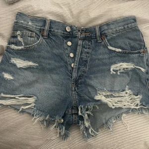 Blå jeansshorts med slitningar och knappar - Säljer ett par blå jeansshorts med råa, fransiga kanter och slitningar framtill. Perfekta för sommaren och en avslappnad stil. 💗