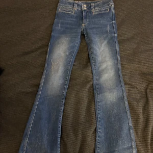 Blå low waist bootcut jeans med dubbla knappar - Snygga blå bootcut jeans med dubbla knappar i midjan och klassiska fickor fram. Jeansen har en lätt tvättad look och utsvängda ben, perfekt för en trendig vardagsstil.