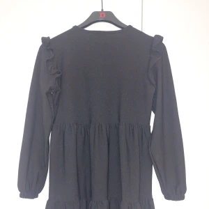 Svart långärmad klänning med volang - En svart klänning med lång ärm och volangdetaljer. Klänningen har en lös passform och är sydd i flera våder för en luftig känsla. Perfekt för dig som gillar en enkel men stilren look.