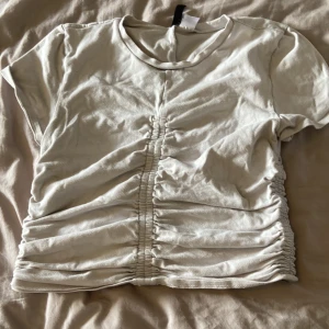 Vit croppad t-shirt med rynkade detaljer - Säljer en vit croppad t-shirt från Bik Bok med rynkade detaljer framtill och baktill. T-shirten har korta ärmar och en rund halsringning. Perfekt för en avslappnad och trendig look. ca 64 cm i midjan