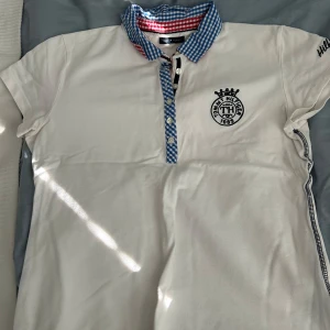 Vit piké från Tommy Hilfiger med rutiga detaljer - Vit pikétröja från Tommy Hilfiger med blå- och vitrutiga detaljer på krage och knappslå. Broderad logga på bröstet och liten svart text på ärmen. Klassisk och stilren design med korta ärmar.