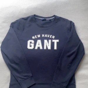 Mörkblå sweatshirt från GANT - Säljer en mörkblå sweatshirt från GANT med vit text 'NEW HAVEN GANT' på bröstet. Tröjan har rund halsringning och långa ärmar. Perfekt för en avslappnad och stilren look.