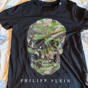 Svart t-shirt med dödskalle från Philipp Plein - Cool svart t-shirt från Philipp Plein med en stor dödskalle framtill i camouflage och glittriga detaljer. T-shirten har rund halsringning och korta ärmar. Perfekt för dig som vill sticka ut med en unik design.