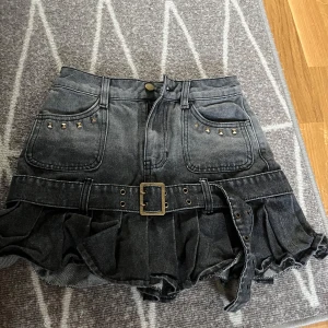 Svart jeanskjol  - Cool svart jeanskjol från Chic Park med volangkant och bälte med spänne. Kjolen har fickor fram med små nitar som detalj och en kort passform. Perfekt för dig som gillar edgy stil.