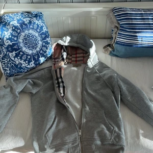 Grå hoodie från Burberry - Säljer en grå hoodie från Burberry med dragkedja och klassiskt rutigt foder i huvan och insidan. Den är XL men har crympt i tvätten till m
