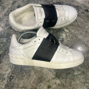 Valentino Open sneakers i vitt och svart - Säljer ett par vita Valentino Open sneakers med bred svart rem över mitten. Skorna har rund tå, platt sula och dekorativa nitar baktill. Klassisk och stilren design med snörning framtill och diskret logga på plösen. Skorna är i 42 och inget of ingår
