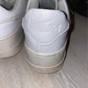 Vita Nike Air Force 1 sneakers - Klassiska vita Nike Air Force 1 sneakers med perforerad tå och snörning. Skorna har en platt sula och är tillverkade i skinn. Perfekta för en stilren och sportig look.