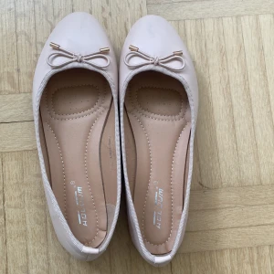Beige ballerinaskor med rosett - Säljer ett par klassiska puderrosa ballerinaskor med söt rosettdetalj framtill och rund tå. Skorna har platt sula och är perfekta för en stilren och enkel look. Passar till både jeans och klänning.
