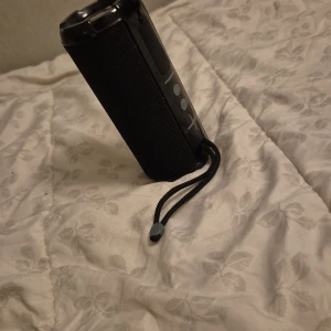 Svart bärbar Bluetooth-högtalare - Säljer en svart bärbar Bluetooth-högtalare med cylinderform och tydliga knappar för volym och play/pause på sidan. Högtalaren har ett praktiskt handtag i tyg för enkel transport. Perfekt för att ta med sig musiken överallt. Kommer med laddare!