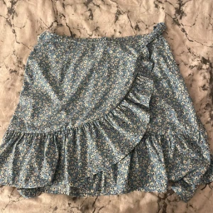 Blommig omlottkjol från ONLY - Söt blå omlottkjol från ONLY med vitt blommönster och volangdetaljer. Kjolen har knytband i midjan och är i en lätt och luftig bomullskvalitet. Perfekt för varma dagar och ger en feminin look.