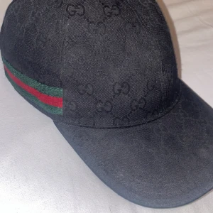 Svart keps från Gucci - Säljer en svart keps från Gucci med diskret GG-monogram över hela kepsen. Klassisk böjd skärm och justerbar rem baktill i svart läder. Sidan har en grön och röd randig detalj som är typisk för Gucci.