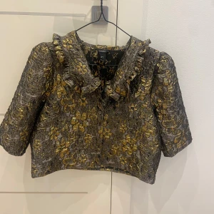 Glittrig kort jacka/kofta med rosett från Lindex - Säljer en glittrig, kort jacka/kofta från Lindex i guld och silver med blommigt mönster. Jackan har trekvartsärm, stor krage och dekorativ rosett framtill. Perfekt för dig som vill sticka ut med en unik och festlig look.