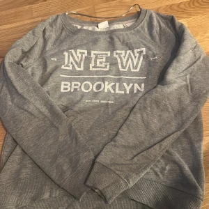 Grå sweatshirt med text från Gina Tricot - Grå sweatshirt från Gina Tricot med vit text 'NEW BROOKLYN' framtill. Tröjan har rund halsringning och långa ärmar. Perfekt för en avslappnad och sportig stil.