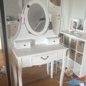 Vit sminkspegel med förvaring - Säljer ett vitt sminkbord med stor oval spegel och tre lådor för förvaring av smink och accessoarer. Bordet har klassisk design med svängda ben och små svarta knoppar på lådorna. Perfekt för att organisera sminkprodukter och tillbehör.