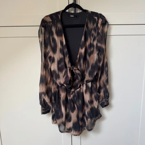 Leopardmönstrad jumpsuit med knyt - Säljer min jumpsuit med leopardmönster i svart och beige toner. Klänningen har djup v-ringning, omlottknytning framtill och resår i midjan för en smickrande passform. 