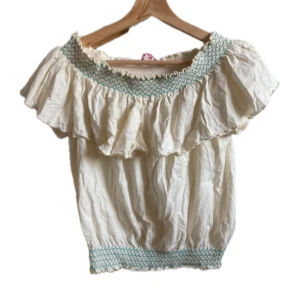 Vit offshoulder blus med volang - Söt vit offshoulder blus med bred volang runt axlarna och smockdetaljer i ljusblått vid halsringning och nederkant. Perfekt för varma dagar och ger en romantisk känsla till outfiten. Den har inga defekter utan är i jättebra skick! 