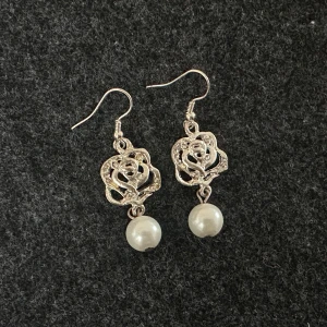 Silvriga örhängen med ros och pärldetalj - Säljer ett par eleganta örhängen i silverfärg med en utskuren ros och en hängande vit pärla. Perfekta för dig som vill ha något unikt och stilrent till din look.