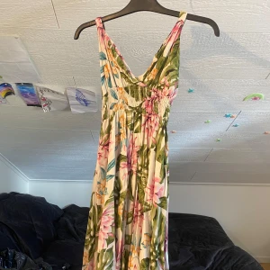 Blommig långklänning med smock - Säljer en somrig långklänning med blommigt mönster i rosa, turkos, orange och grönt. Klänningen har breda axelband, smock upptill och v-ringad halsringning. Perfekt för varma dagar och midsommarfirande.