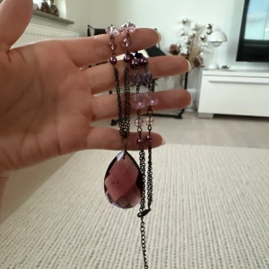 Långt halsband med lila och rosa pärlor - Säljer ett långt halsband med en stor, droppformad lila sten som hänge. Kedjan är dekorerad med pärlor i lila, rosa och blå nyanser. Perfekt för att ge din outfit en färgklick och unik touch.
