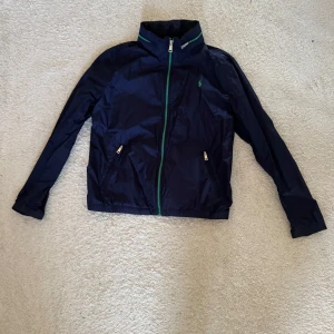 Polo Ralph Lauren vindjacka mörkblå - Snygg mörkblå vindjacka från Polo Ralph Lauren med gröna detaljer och dragkedja framtill. Jackan har ståkrage, två fickor med dragkedja och klassisk logga broderad på bröstet. Perfekt för svala dagar. Jackan har även en luva som sitter i kragen. 