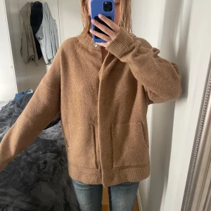 Beige stickad kofta med fickor - Mysig beige stickad kofta med två stora fickor framtill och diskreta knappar. Koftan har lång ärm och en rak, loose passform. Perfekt att slänga på sig över en t-shirt eller topp för extra värme.